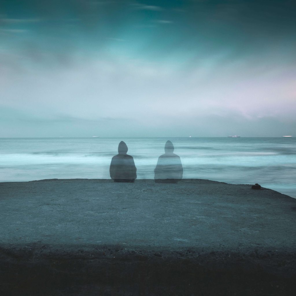 Adult looking at the ocean on a cliff in a moody setting, with a shadow version of themself sat beside them representing loneliness.